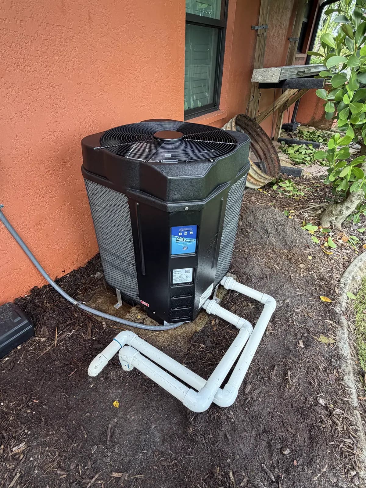 GulfStream HE110-RA pool heat pump beside an orange stucco wall with new white PVC plumbing in a mulch bed
