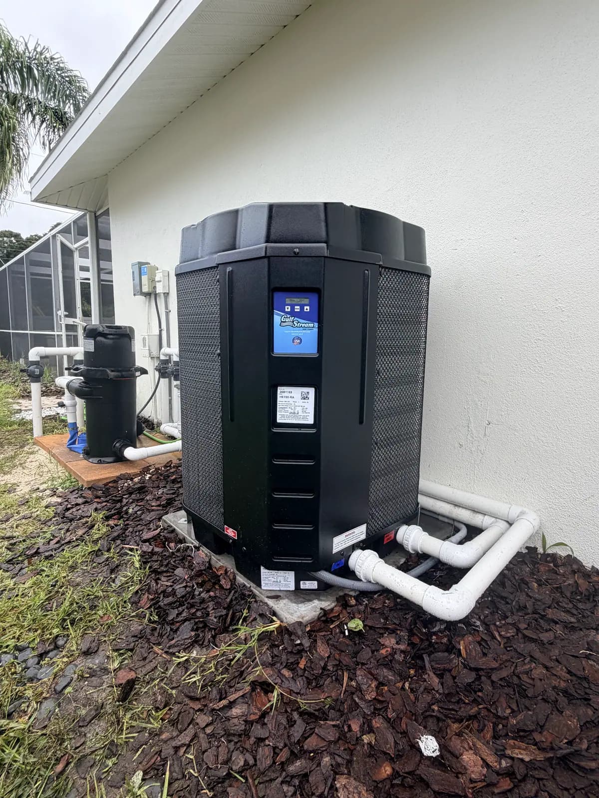 GulfStream HE150-RA pool heat pump on a pad beside a white house wall, with PVC plumbing running along the wall