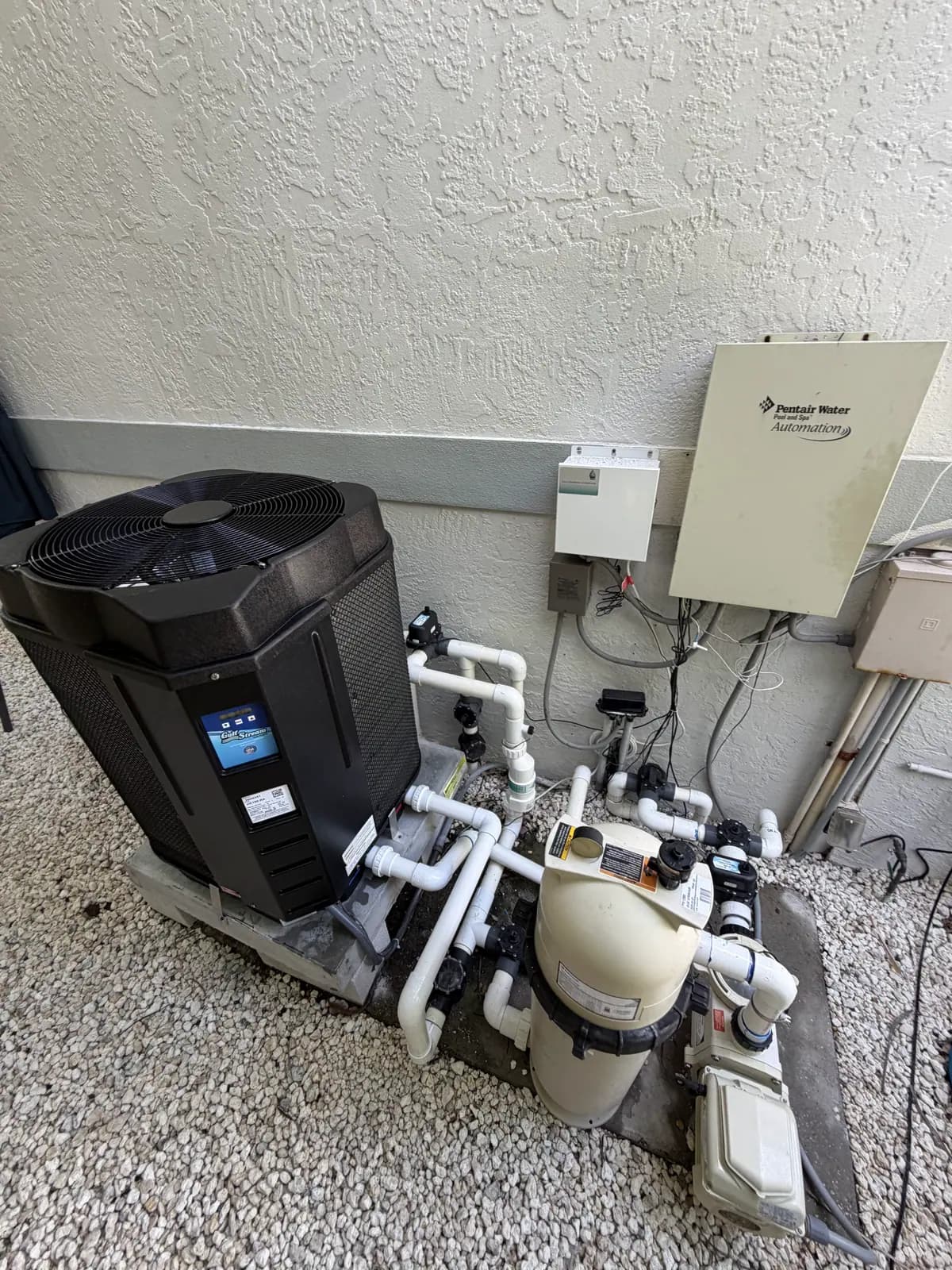 GulfStream heat pump beside a pool filter and pump on white gravel, with a Pentair automation panel on the wall above