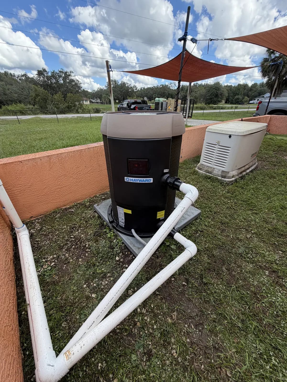 Hayward pool heat pump on an equipment pad in a walled yard with long white PVC pipe runs under shade sails
