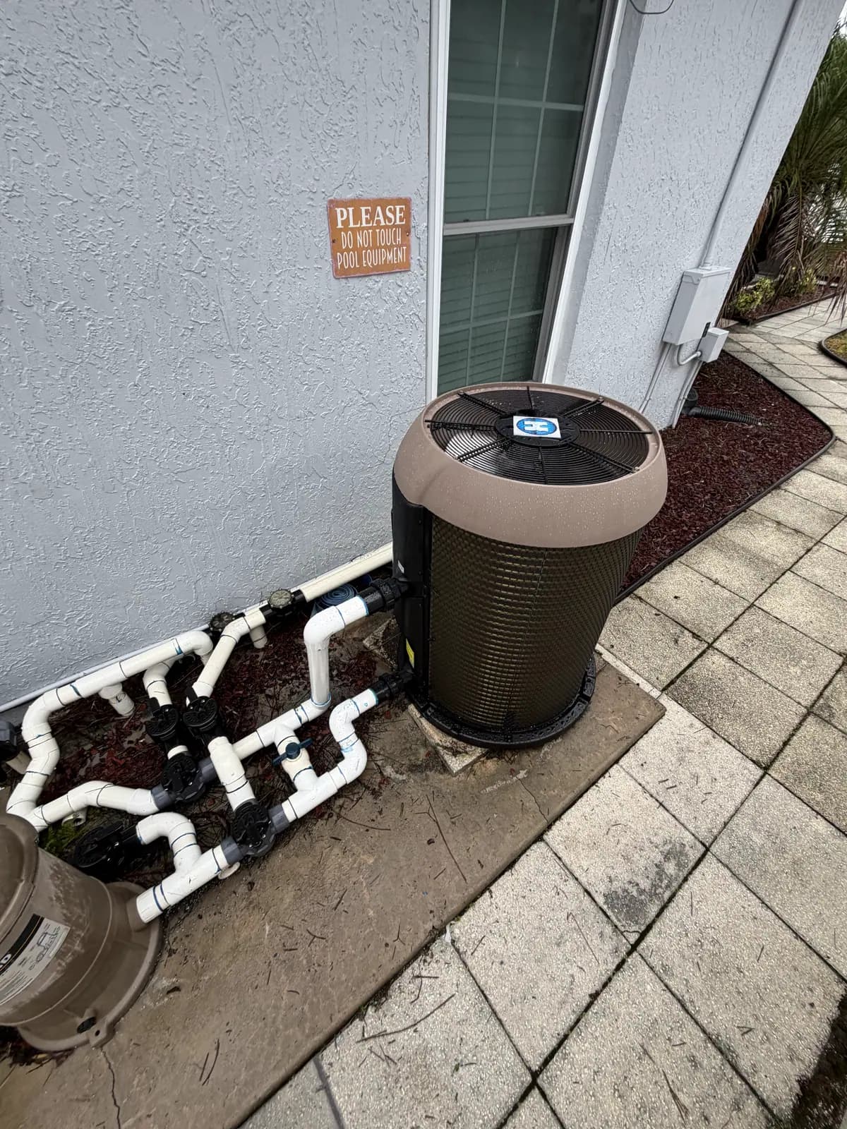 Hayward pool heat pump on a concrete pad beside a gray house wall with a PVC valve manifold and a paver walkway