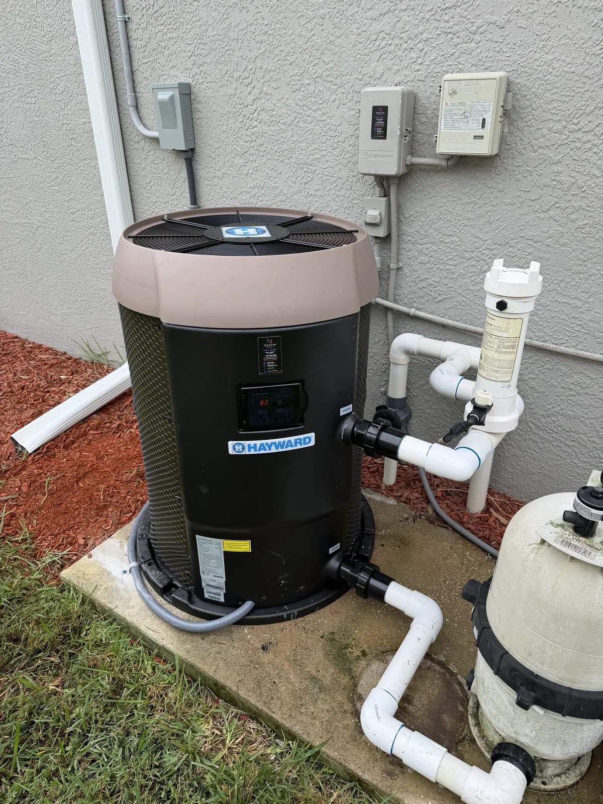 Hayward pool heat pump on a concrete slab beside a pool filter, with PVC plumbing and wall-mounted electrical boxes