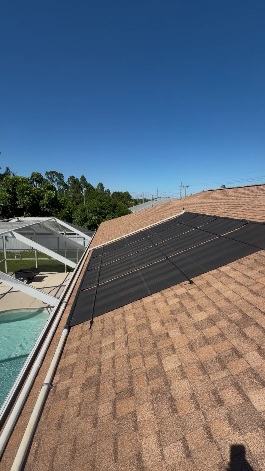 Black solar pool heating collectors on a tan shingle roof beside a screened pool enclosure, under a clear blue sky